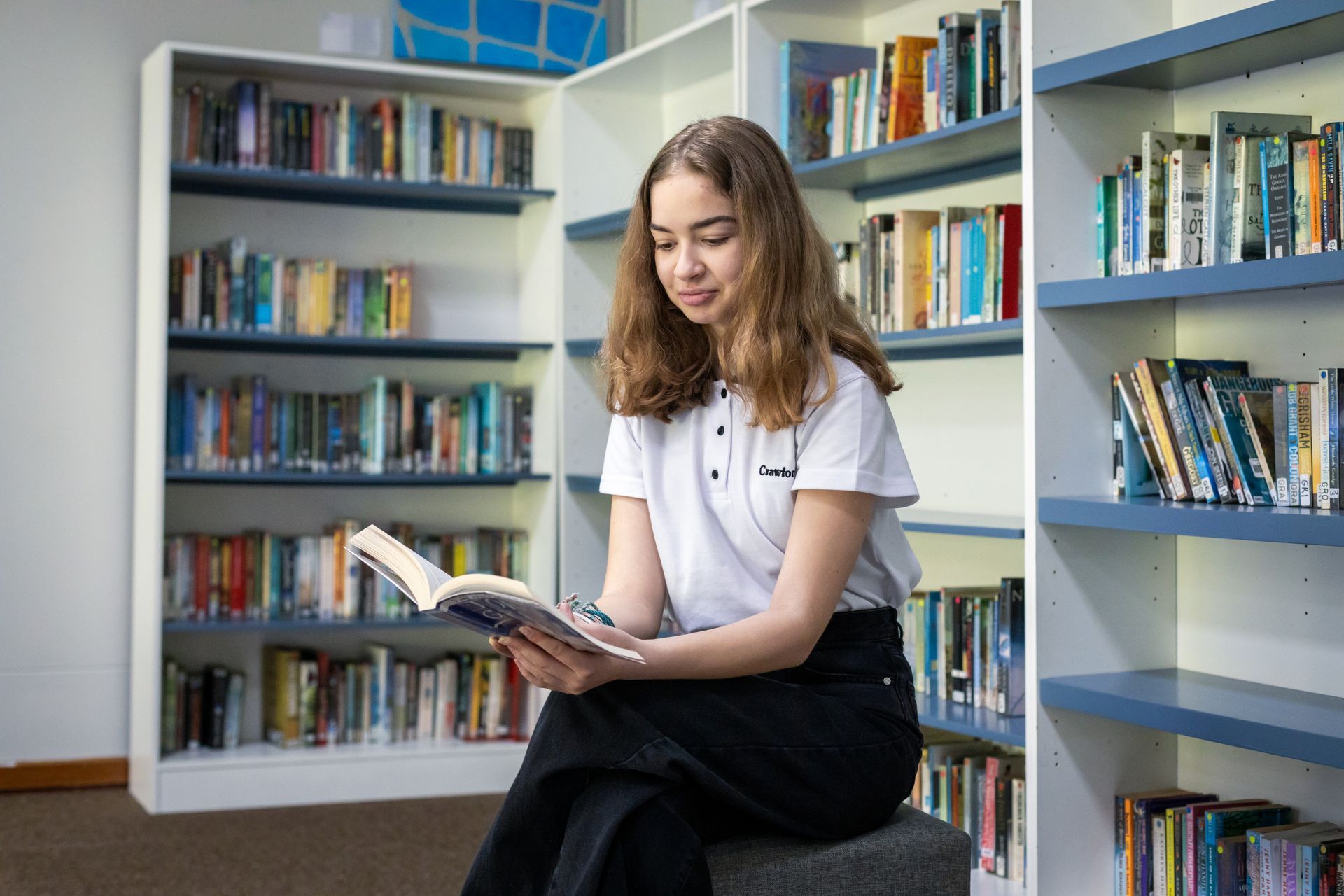 A young girl is sitting on a stool in a library reading a book.