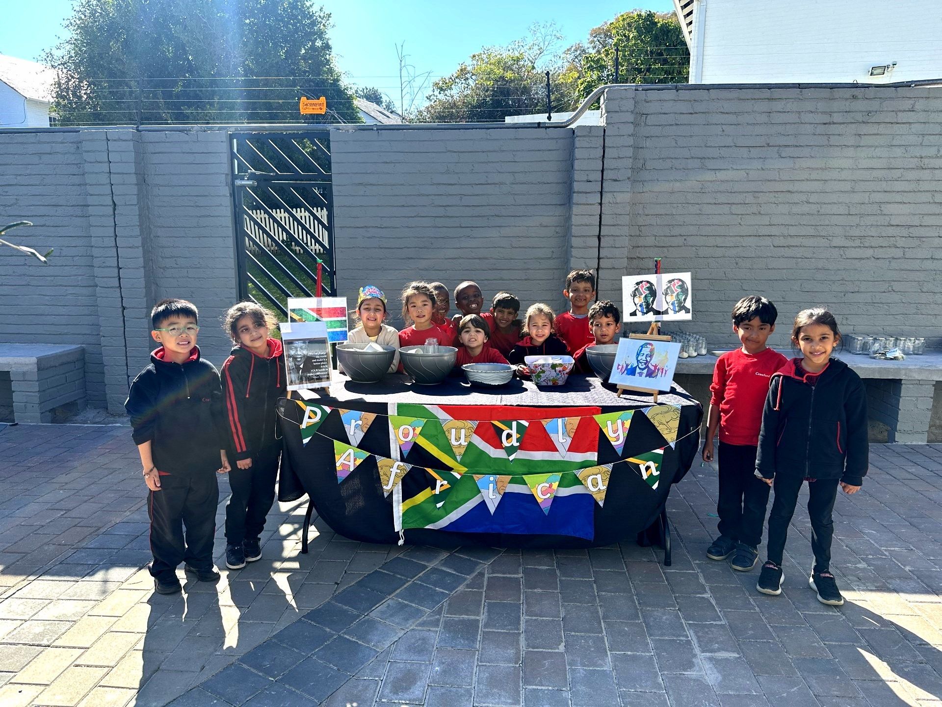 Children at a table with food, decorated with a South African flag, outdoors on a sunny day.