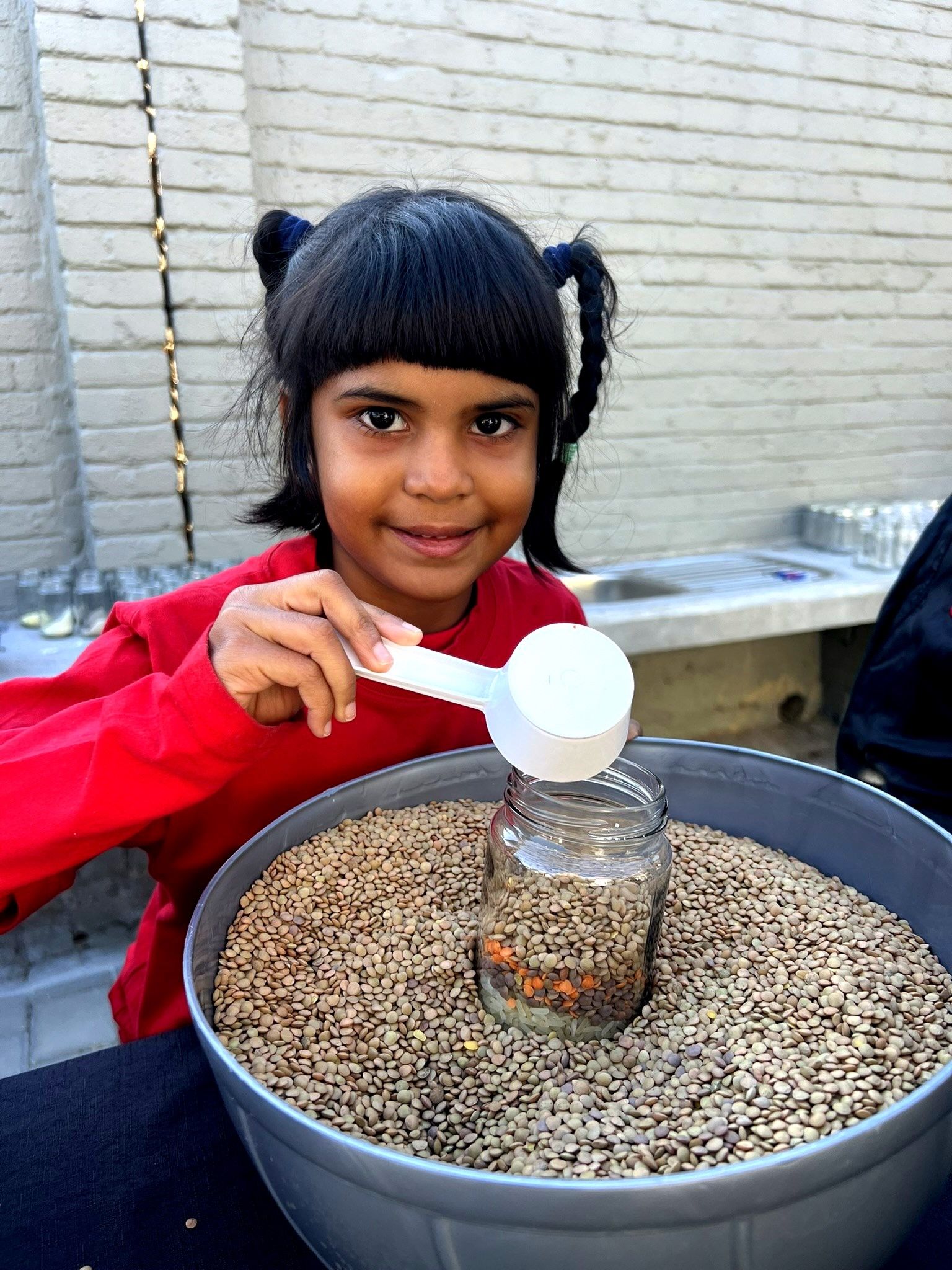 Girl scoops seeds into a jar in a large bowl. She smiles while wearing a red shirt. Brick wall background.