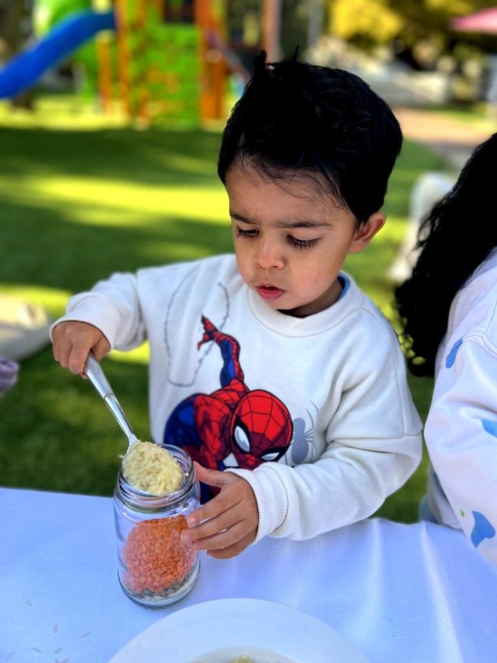 Young child in a Spider-Man shirt using a spoon to add yellow food to a jar on a table outdoors.