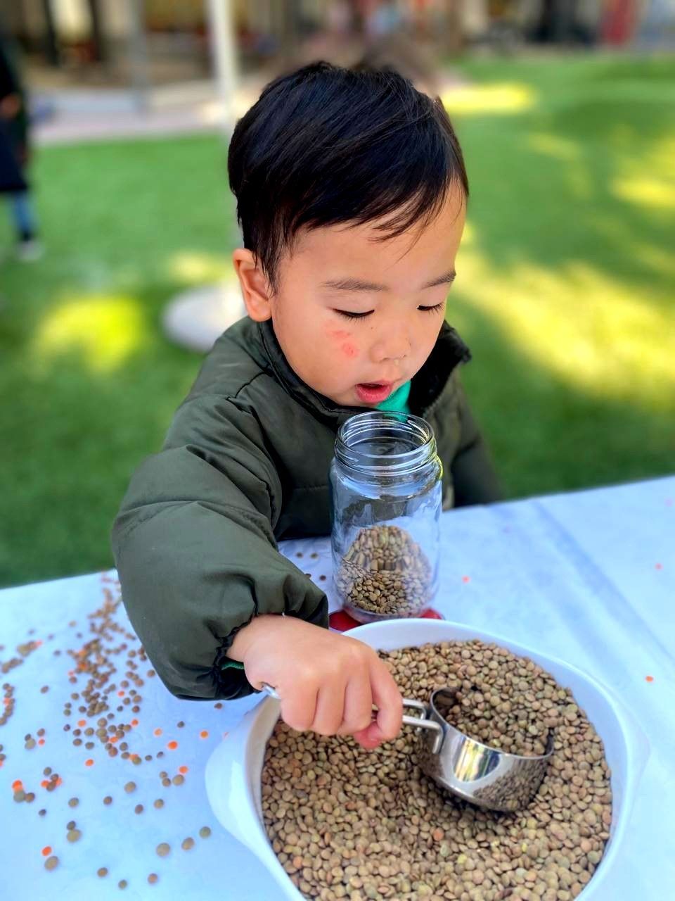 Child with dark hair using a scoop to fill a jar with lentils, outside on a table.
