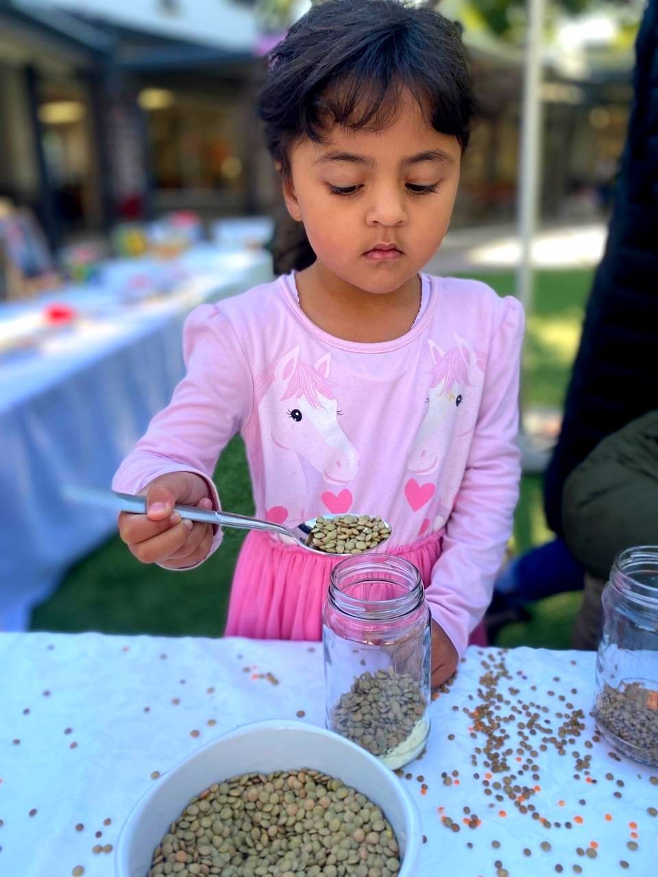 Girl in pink shirt filling a jar with lentils from a spoon at a table.