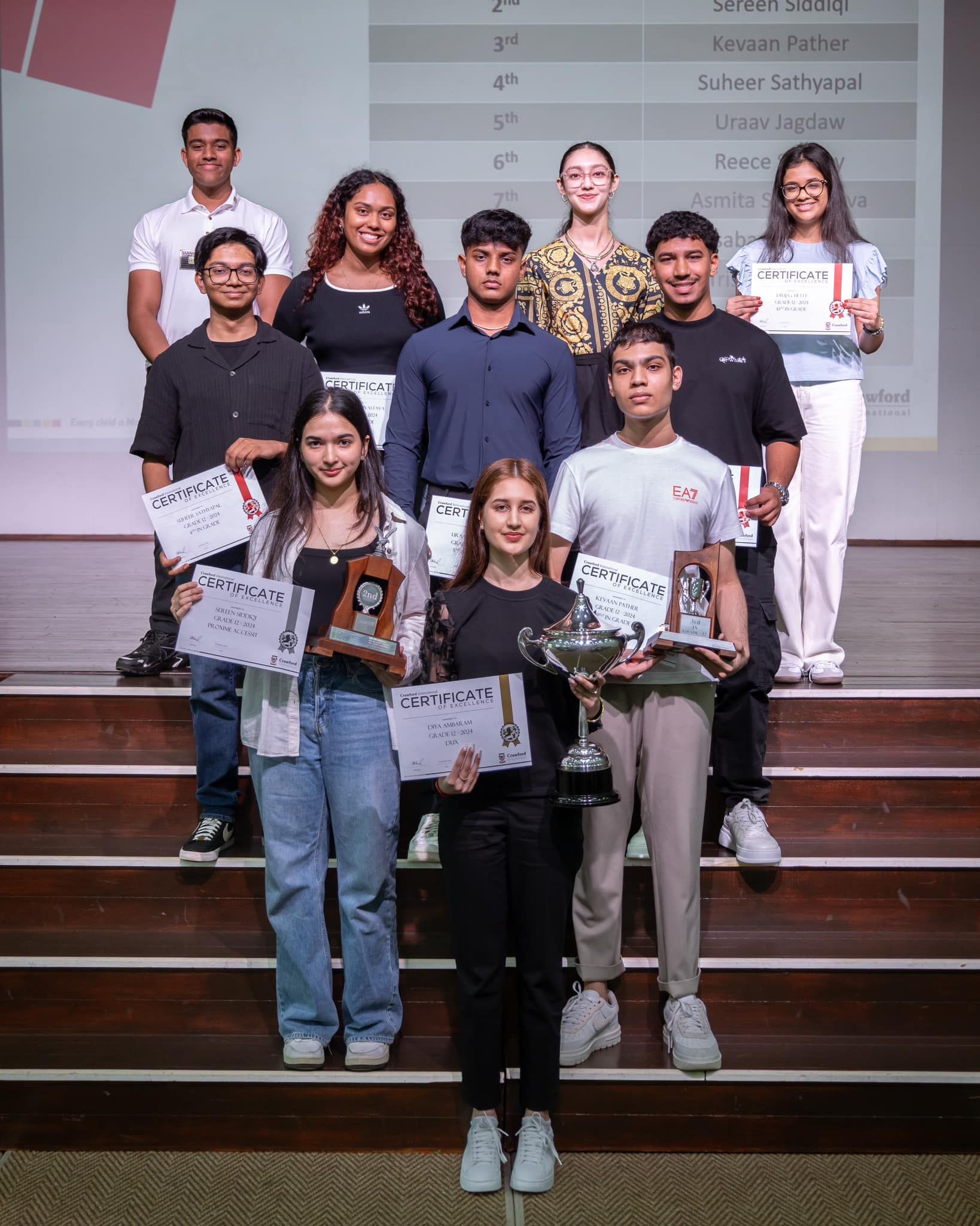 Group of students holding awards, smiling on stage, with a background table.