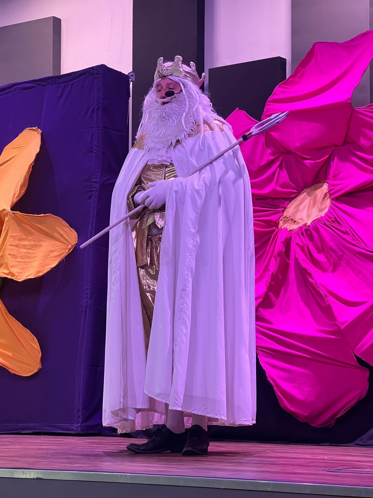 A person in a white king costume holding a scepter on a stage with flower decorations.