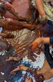 People's legs and arms covered in paint, sitting on a plastic sheet, messy and colorful.