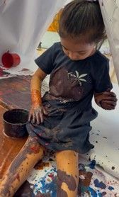 Child covered in brown paint, sitting on a white sheet, appears to be painting.
