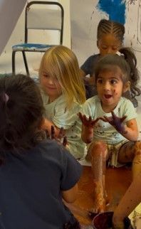 Children painting with hands on floor. Paint is red/brown. Some wearing blue shirts, some with paint on them.