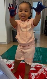 Child in pink outfit with painted hands stands over a paint-splattered surface, smiling with arms raised.