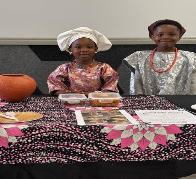 Two children in traditional attire at a table with food, display and a decorative cloth.
