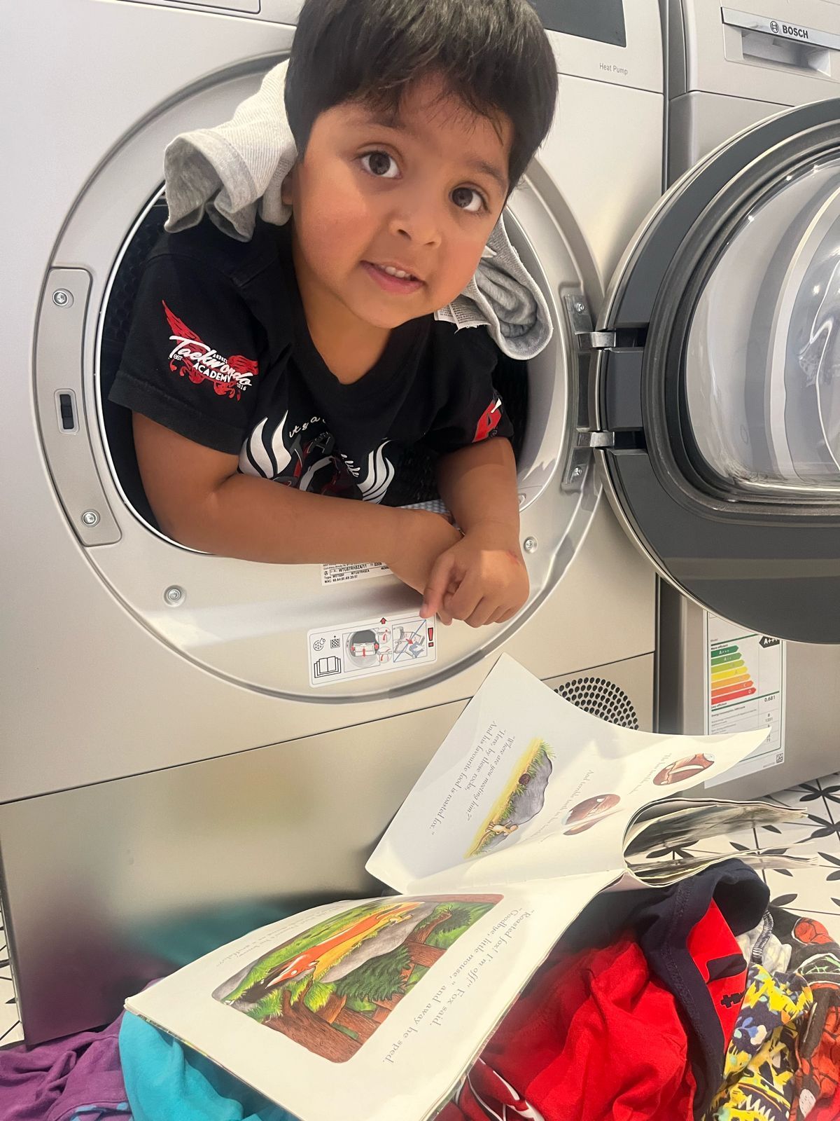 A young boy is sticking his head out of a washing machine while reading a book.