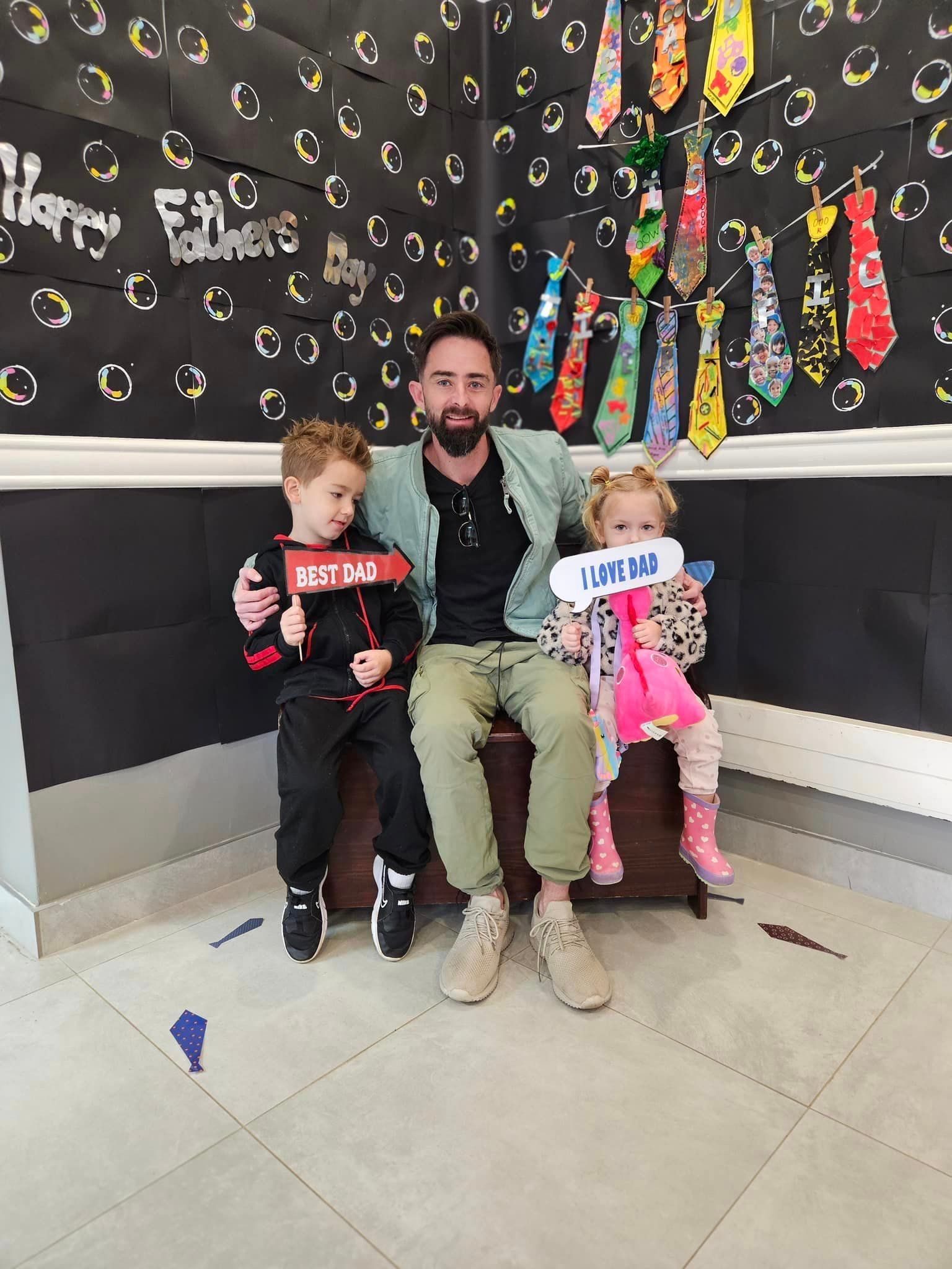 A man is sitting on a stool with two children holding signs.