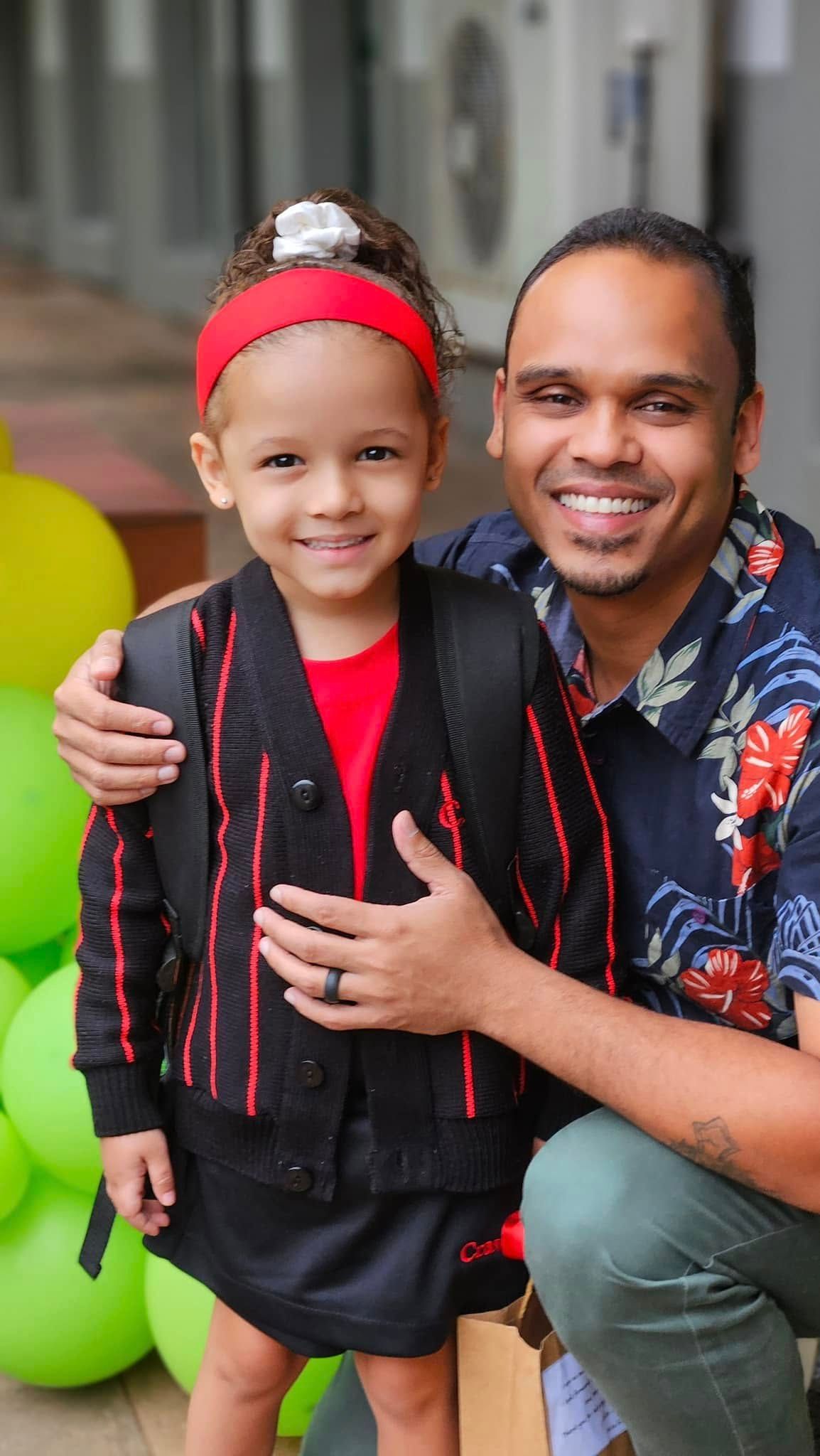 Man and child smiling, embracing outdoors. Child wears red headband and backpack, man has floral shirt.