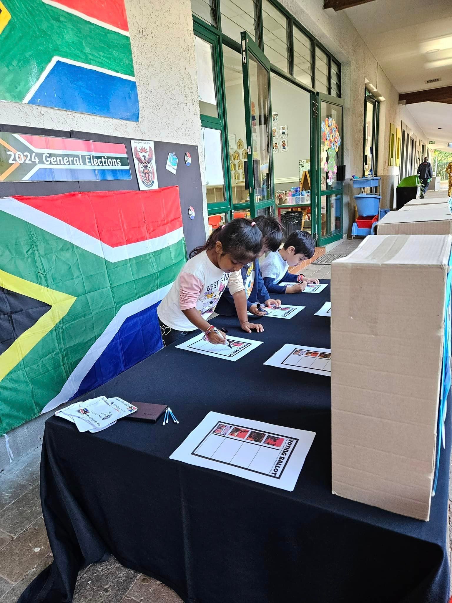 A group of children are sitting at tables in front of a south african flag.