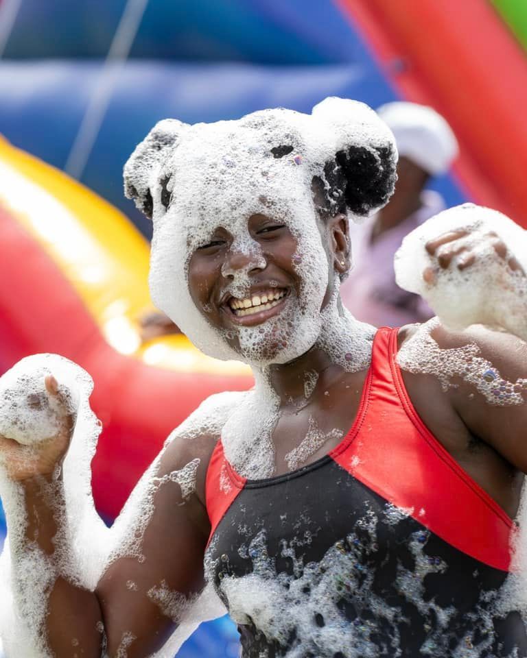 Woman covered in foam, smiling, at a colorful outdoor event.
