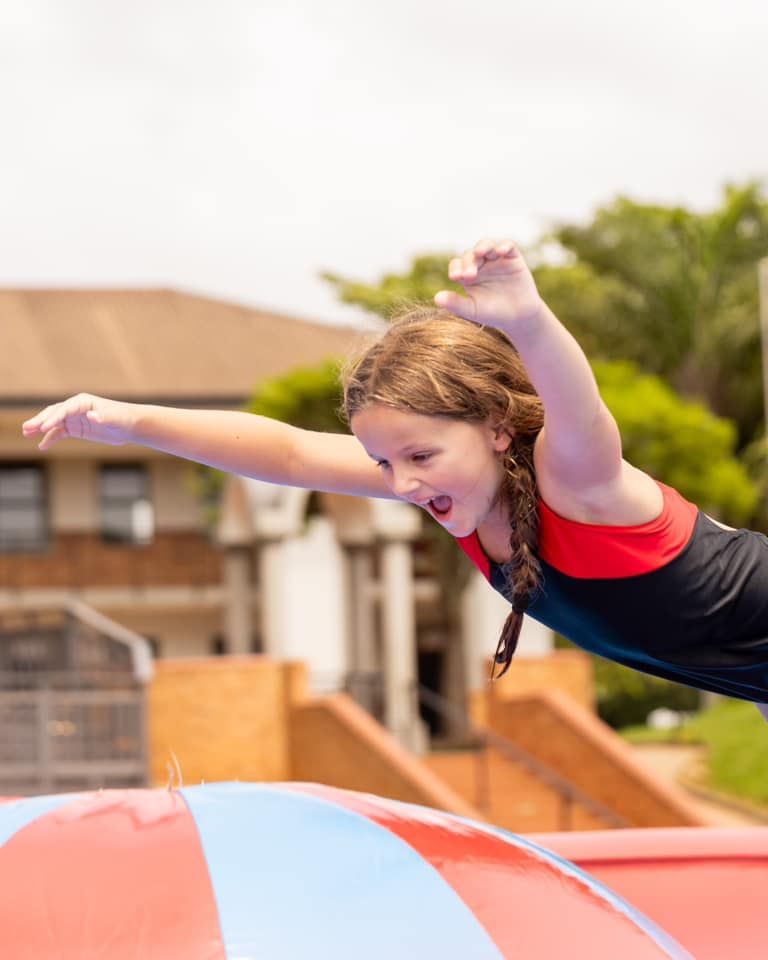 Girl dives through the air, mouth open. Leaping off a red and blue inflatable.