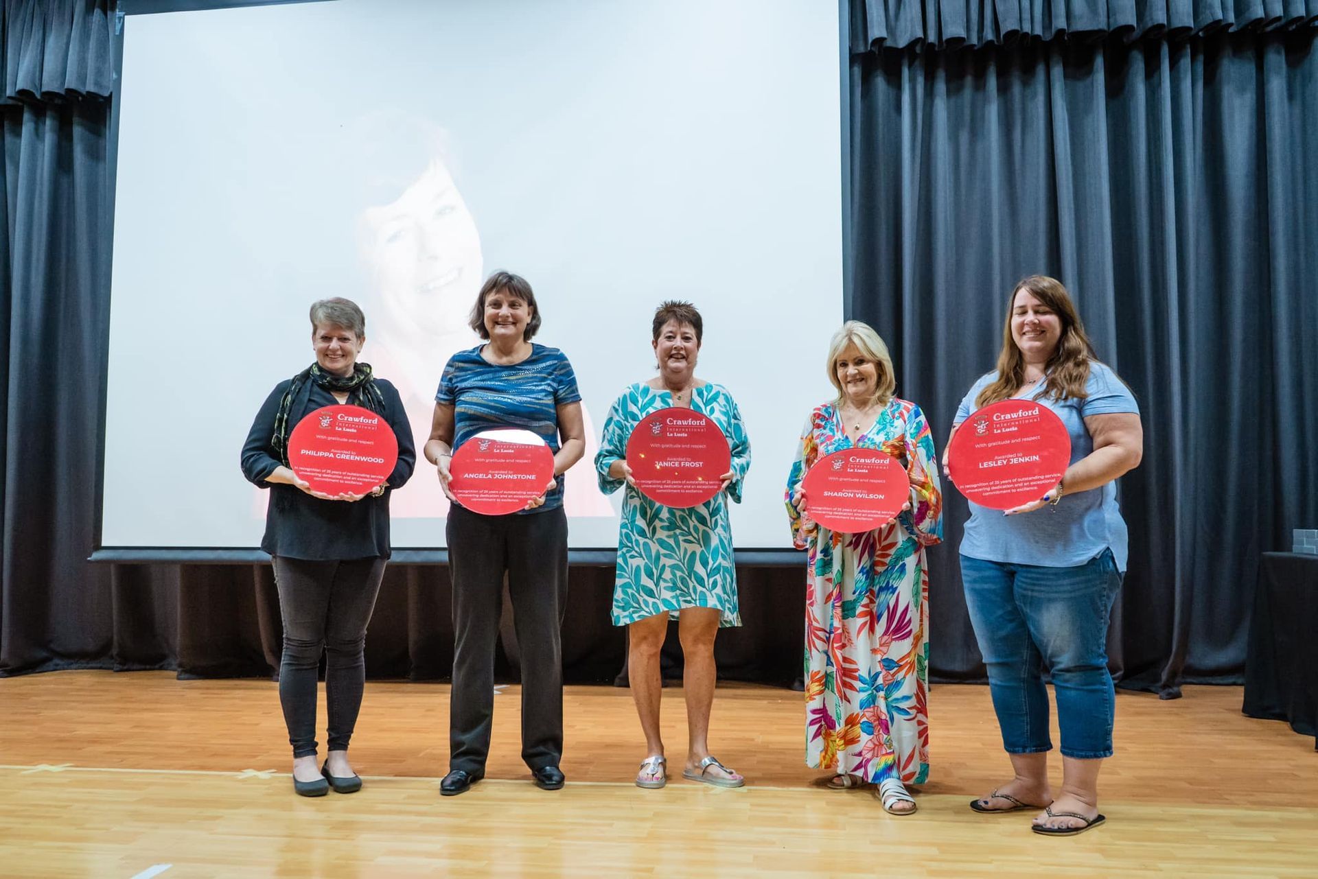 Five women stand holding red circles, smiles, in front of a screen and curtain.