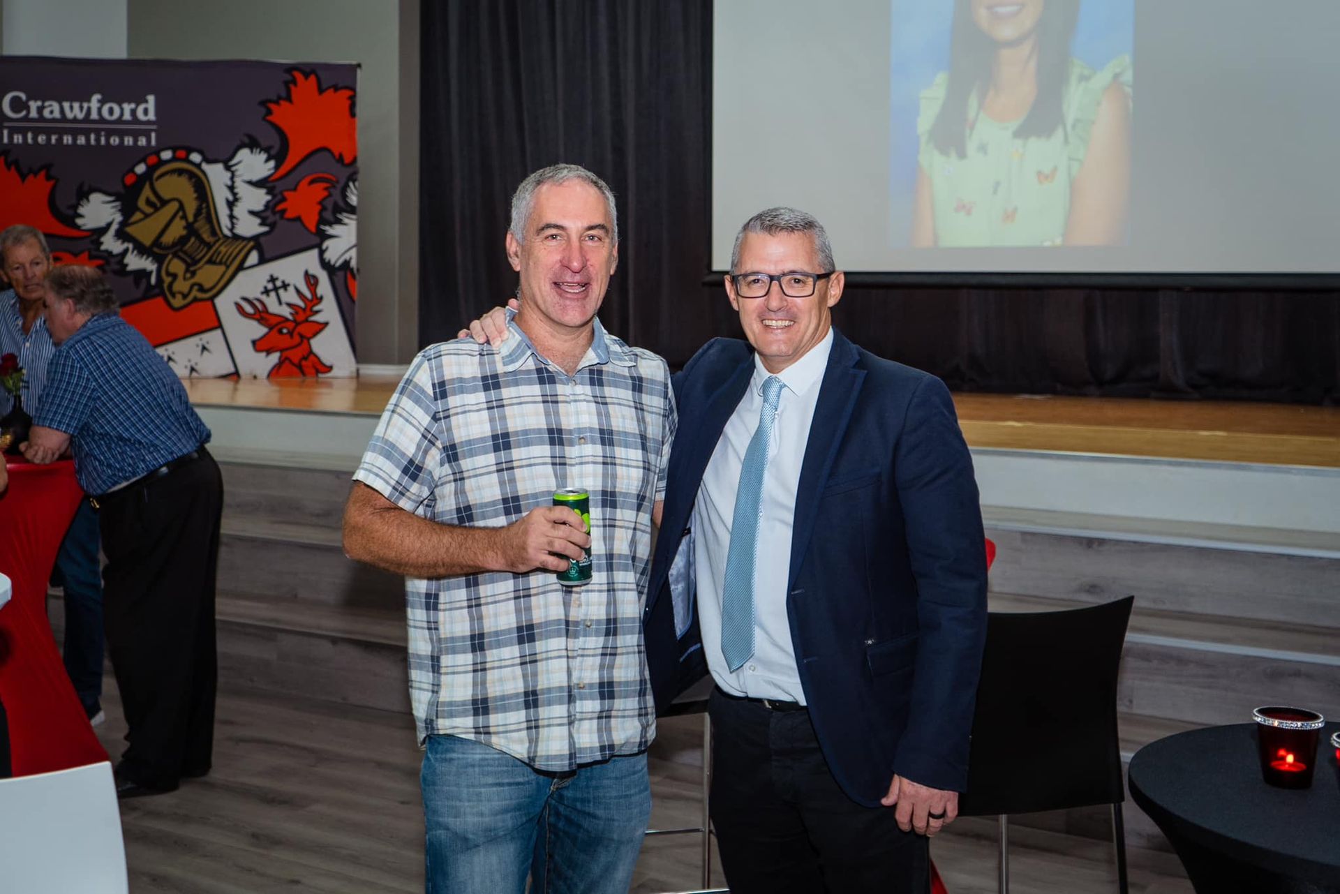 Two men standing, one in a plaid shirt and jeans holding a drink, the other in a suit. They are indoors.
