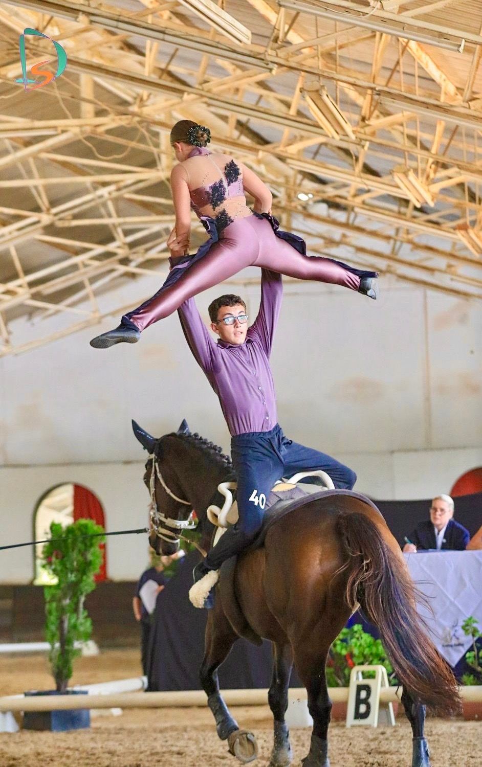 Vaulting on horseback. Man lifts woman in a purple leotard. Indoor arena with judge and audience.