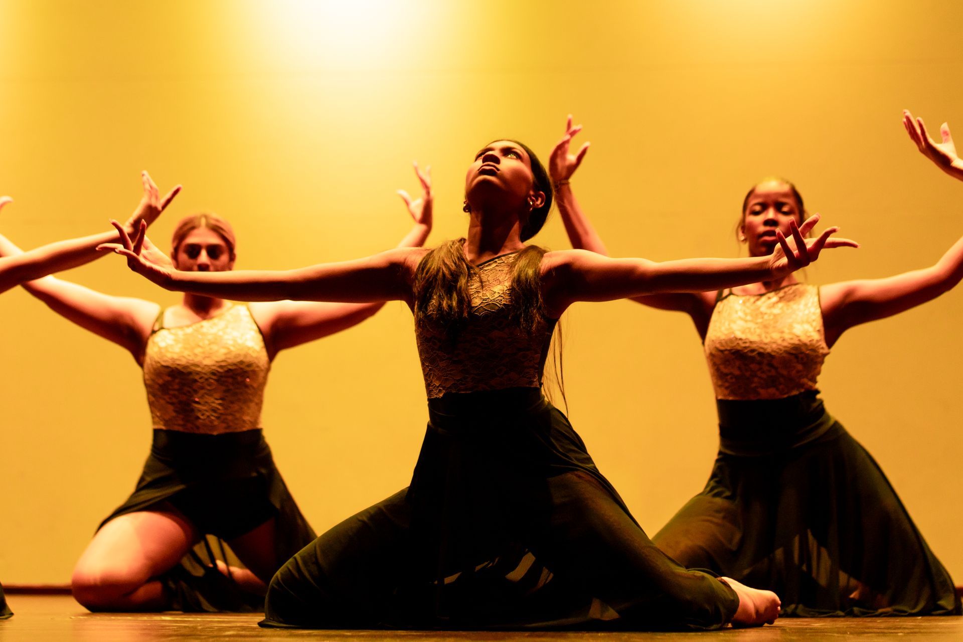 Dancers in gold tops and black skirts performing on a stage with a warm-toned backdrop, arms raised.