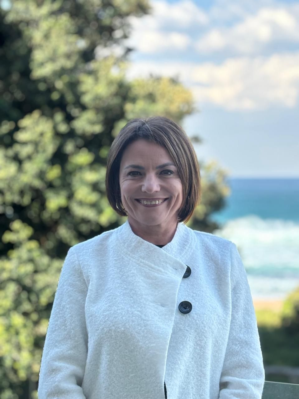 A woman in a white coat is smiling in front of the ocean.