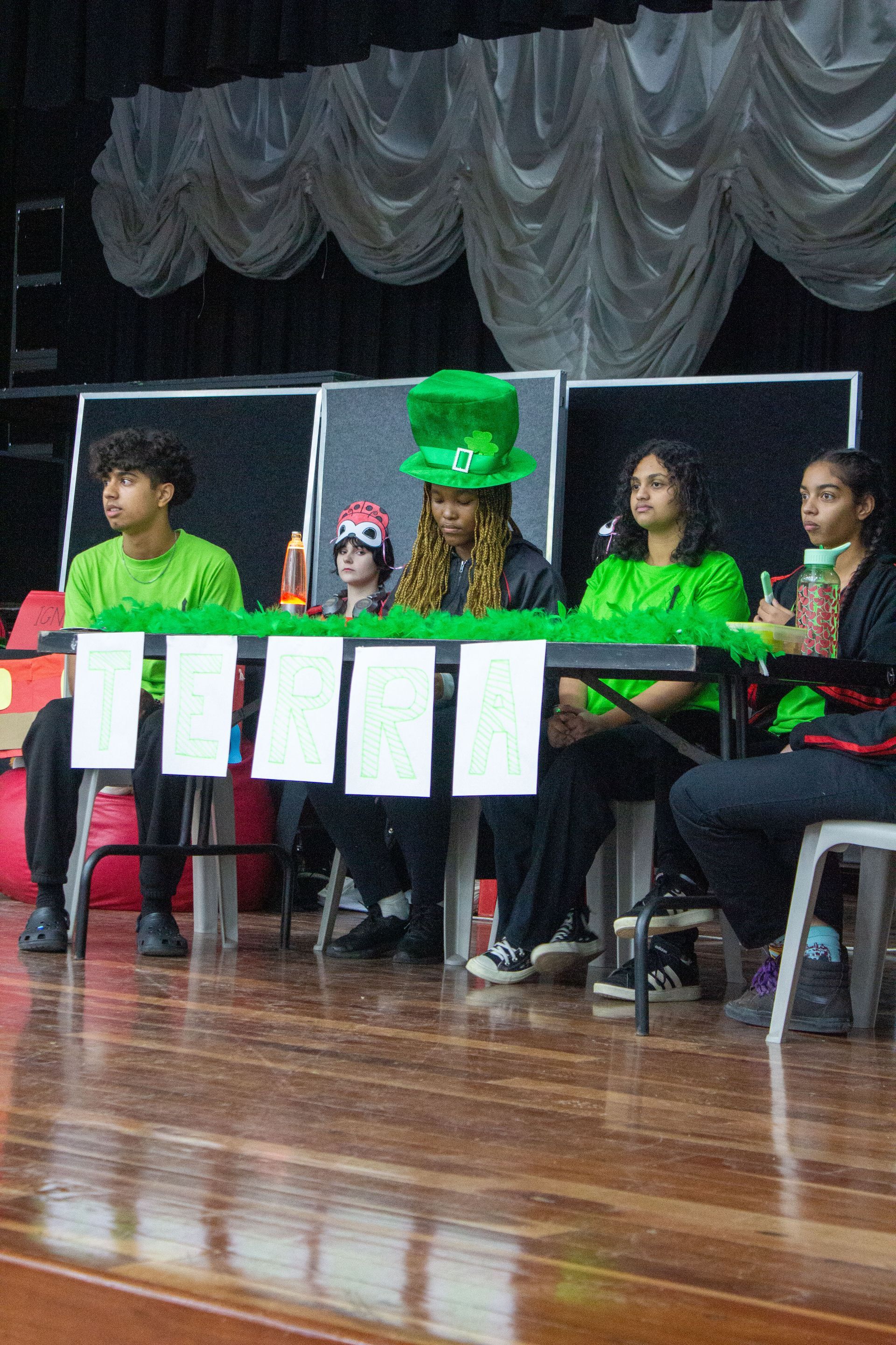 A group of people are sitting at a table on a stage . one of the people is wearing a leprechaun hat.