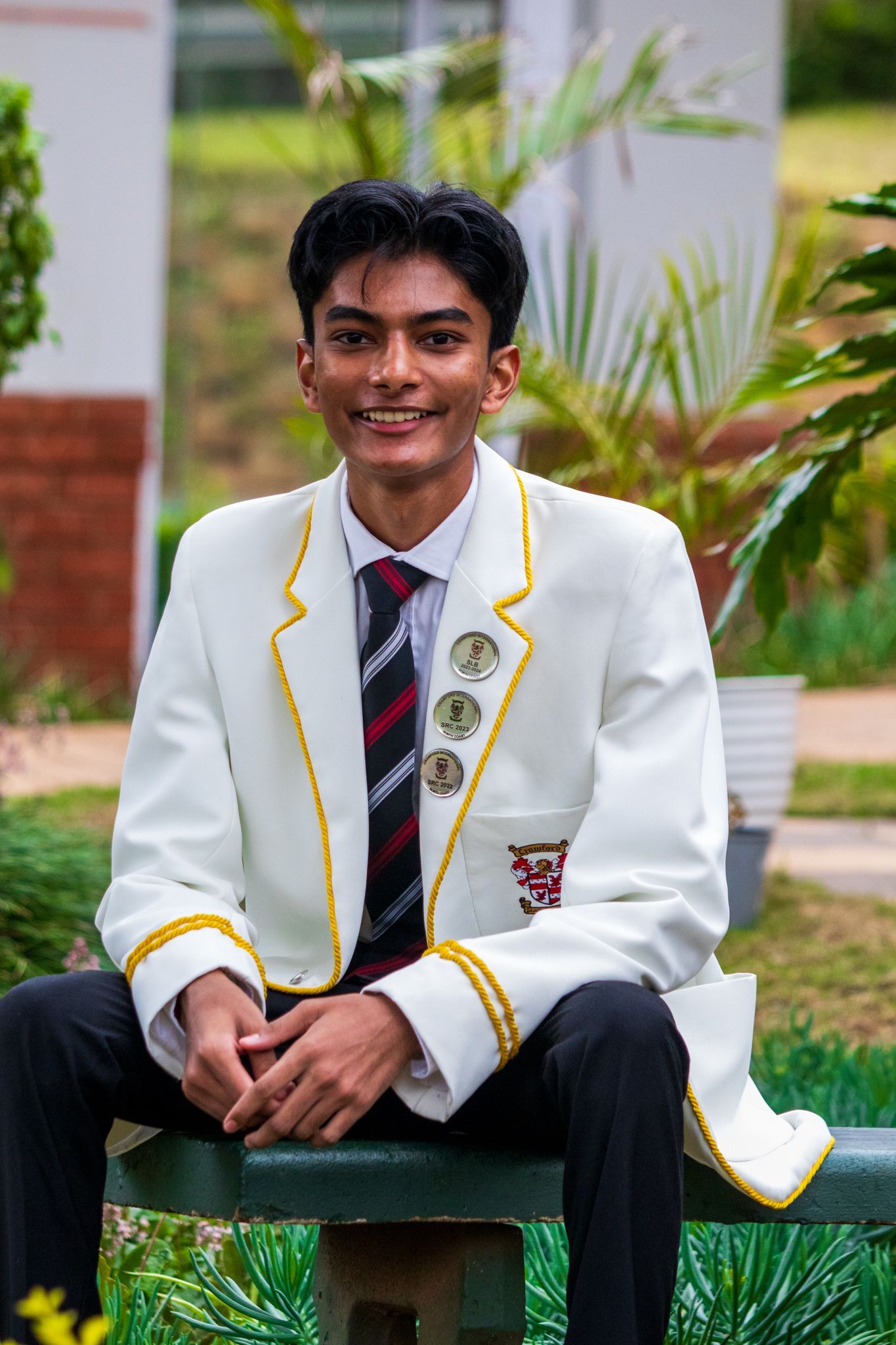 A young man in a white jacket and tie is sitting on a bench.