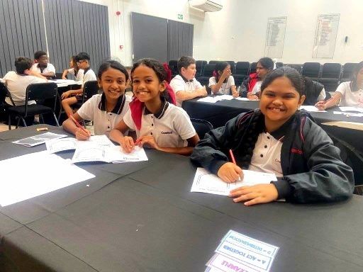 Three smiling students in school uniforms work at a table. Others are in the background, writing.