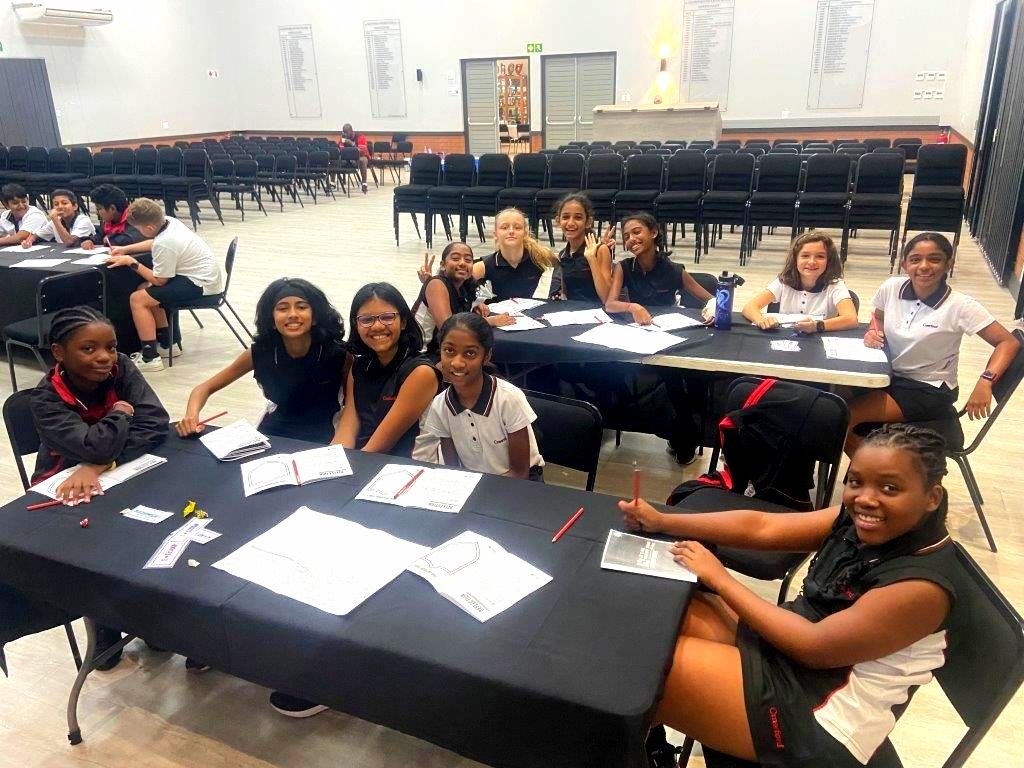 Students at tables in a large room, some smiling, with papers, pens; chairs behind.