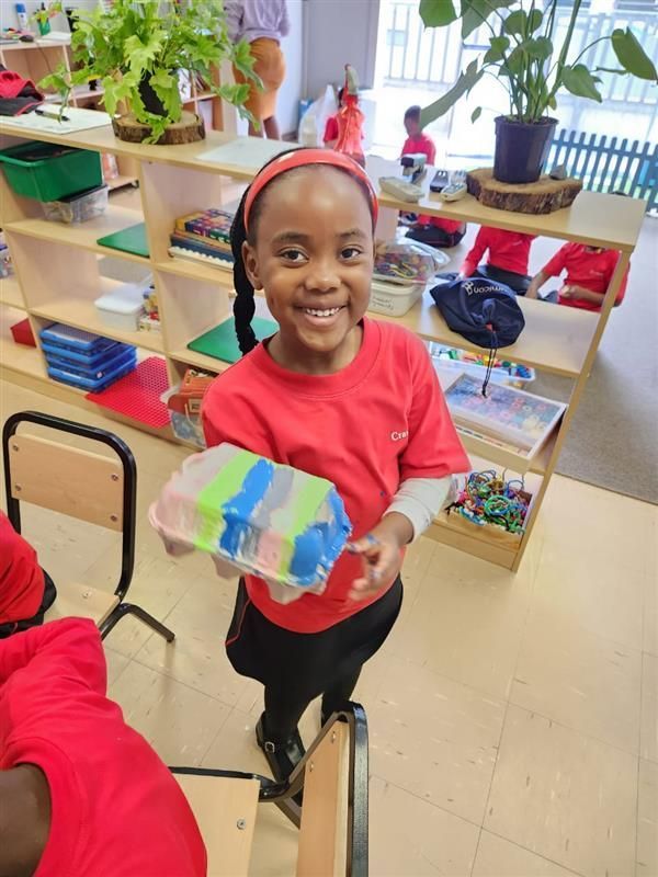 Young child in a red shirt smiles, holding a painted craft in a classroom setting.