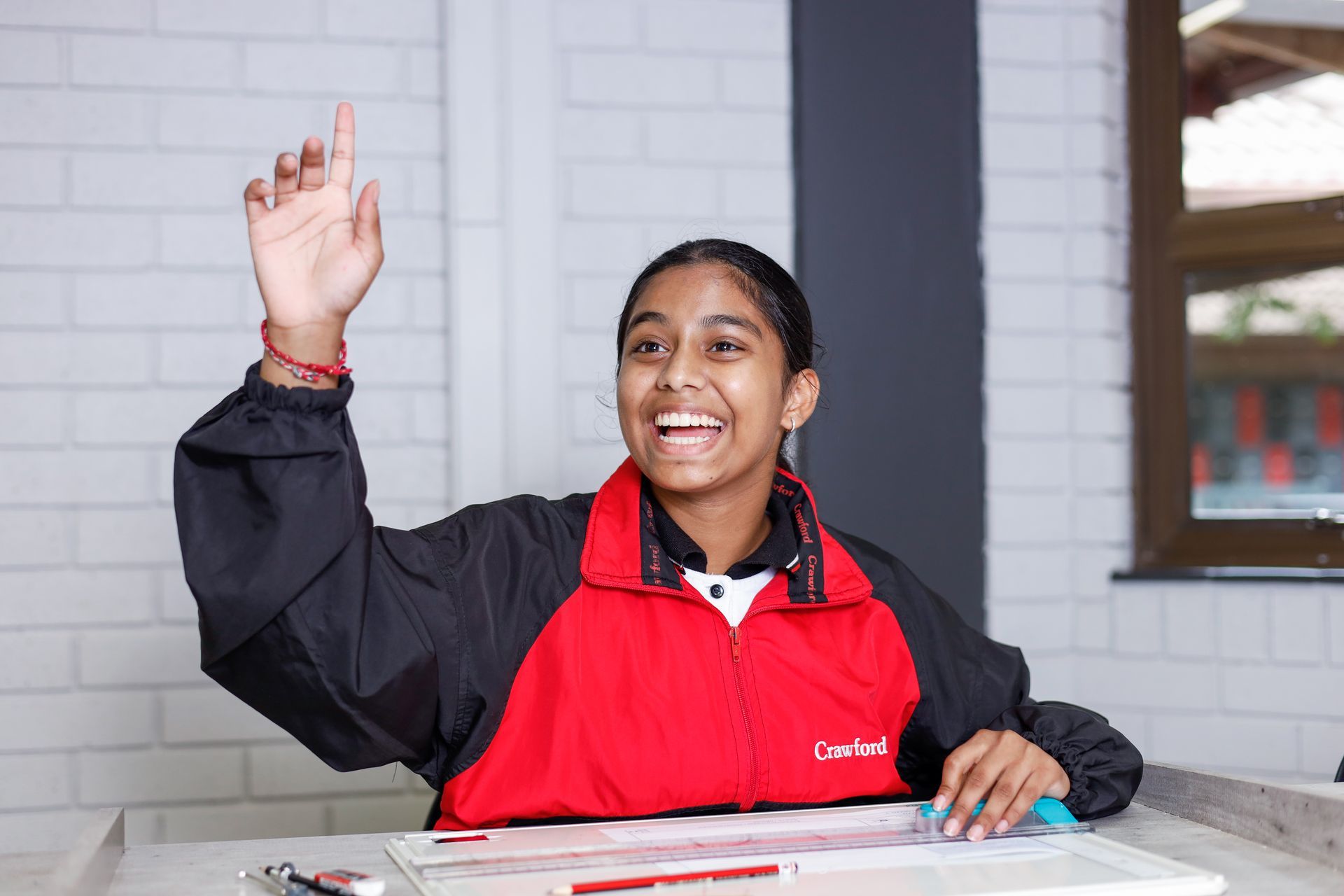 A young girl is sitting at a desk with her hand up.