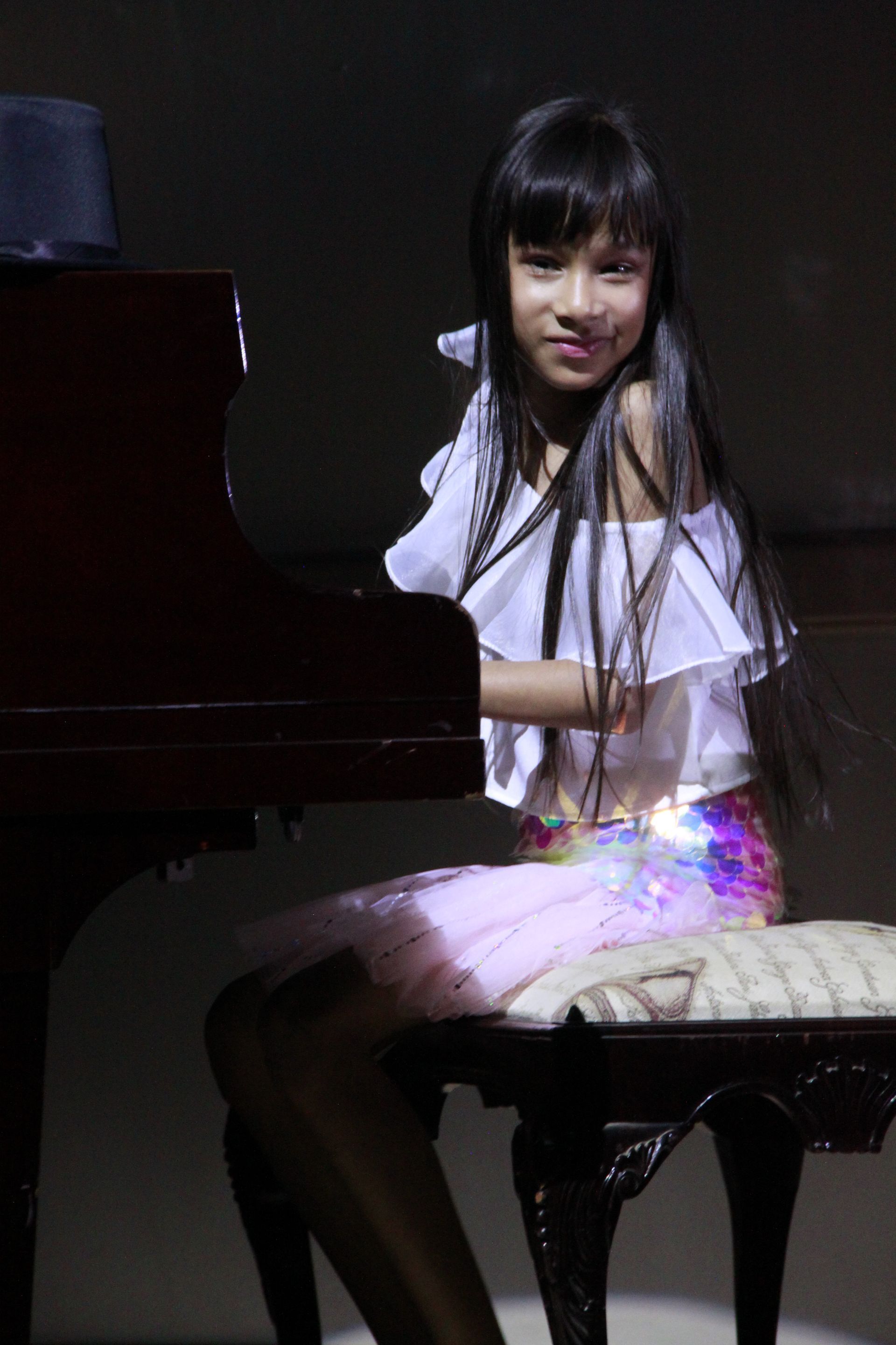 A young girl is sitting on a stool in front of a piano.