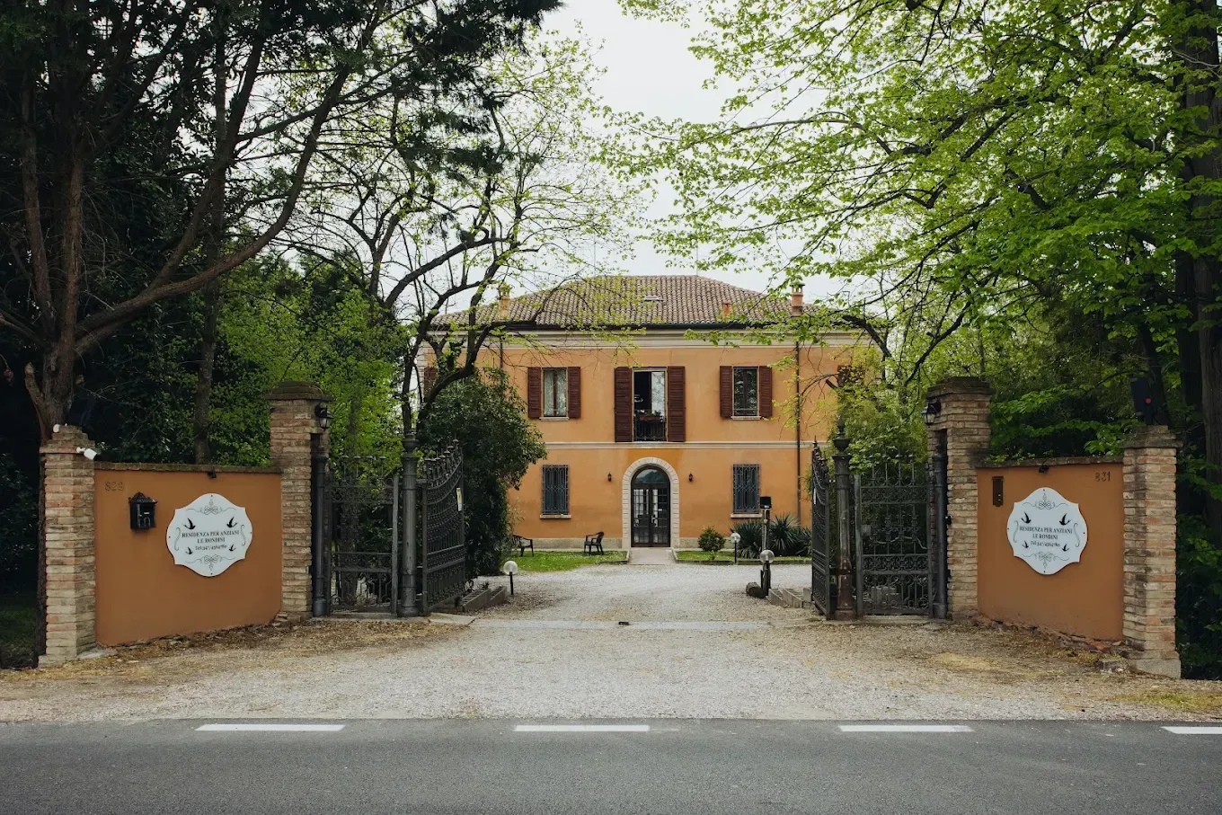 Casa beige con cancelli aperti, alberi e vialetto di ghiaia.