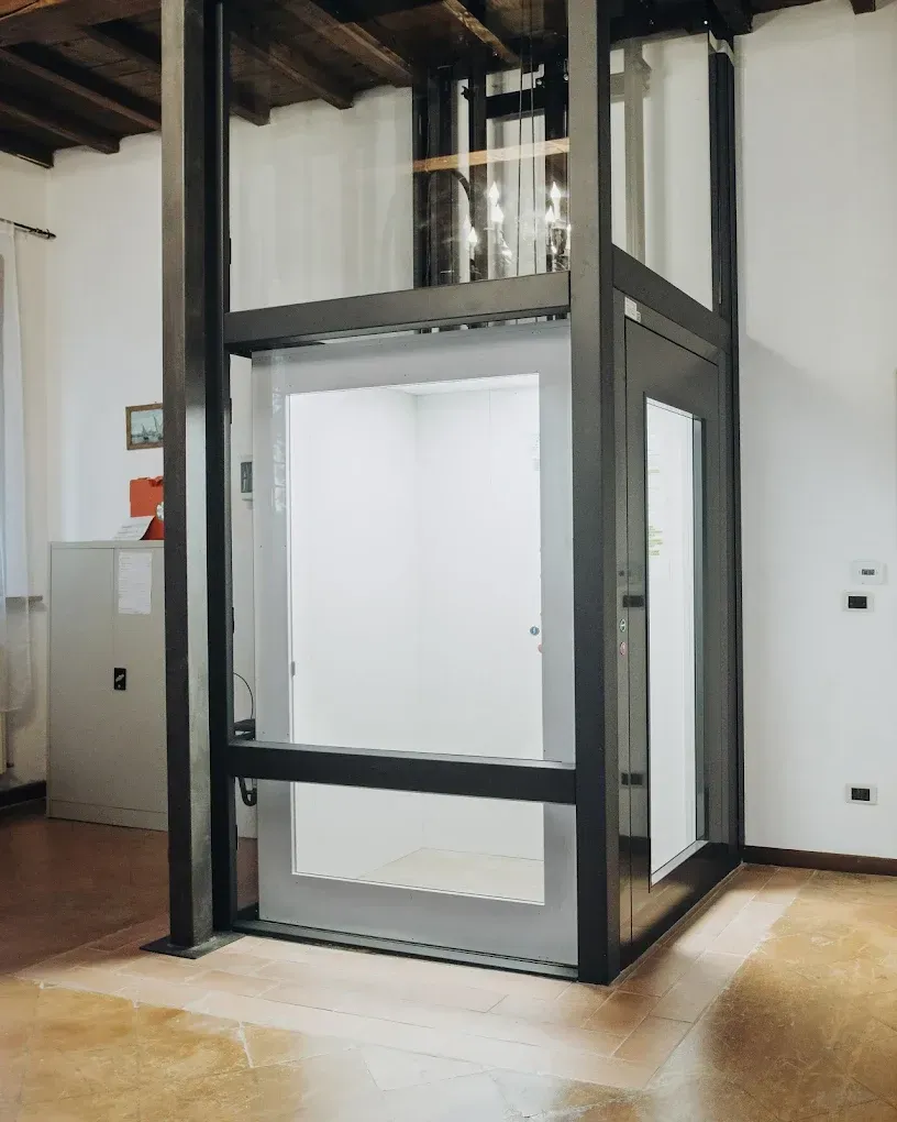 Ascensore chiuso con pareti in vetro e struttura in metallo scuro all'interno di una casa. L'interno è bianco.