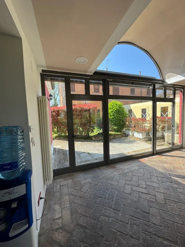 Vista interna di una porta a vetri che conduce a un cortile con cespugli rossi, pavimento in mattoni e refrigeratore d'acqua.