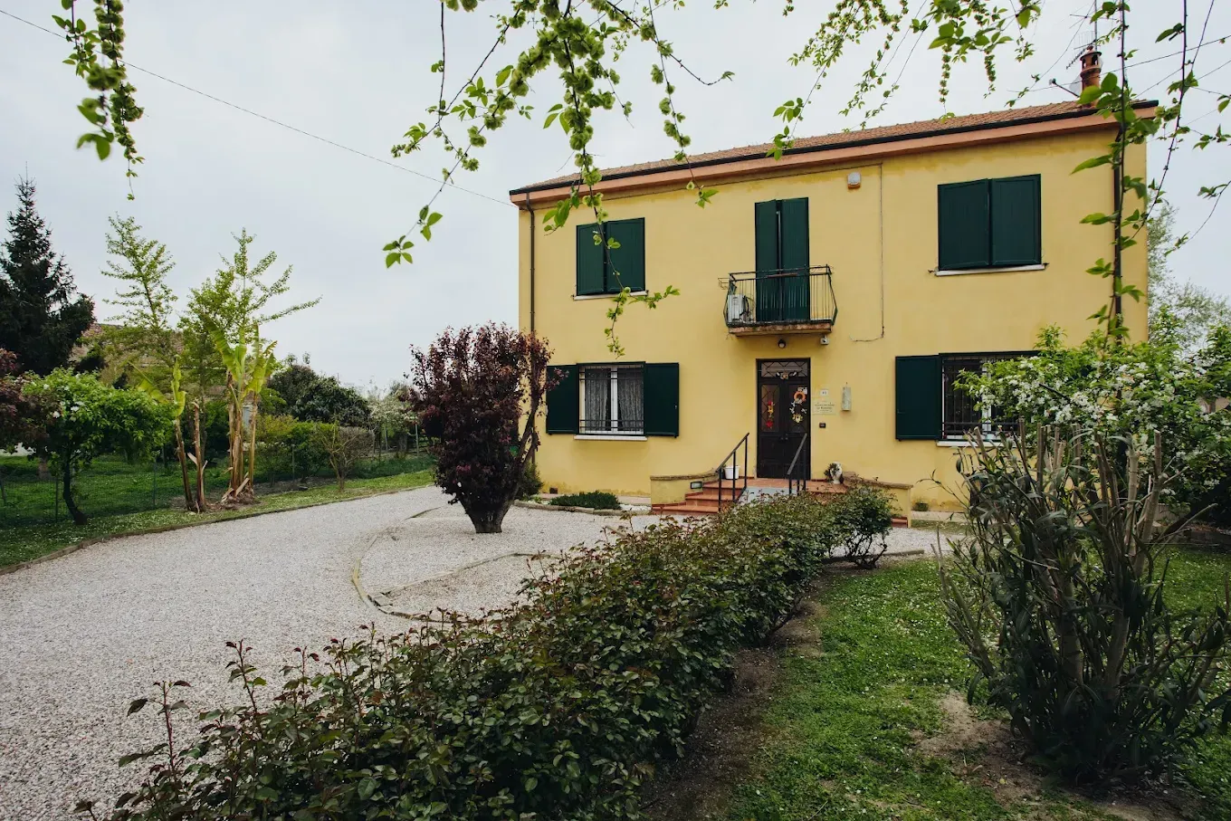Casa gialla a due piani con persiane verdi, vialetto di ghiaia e verde circostante.