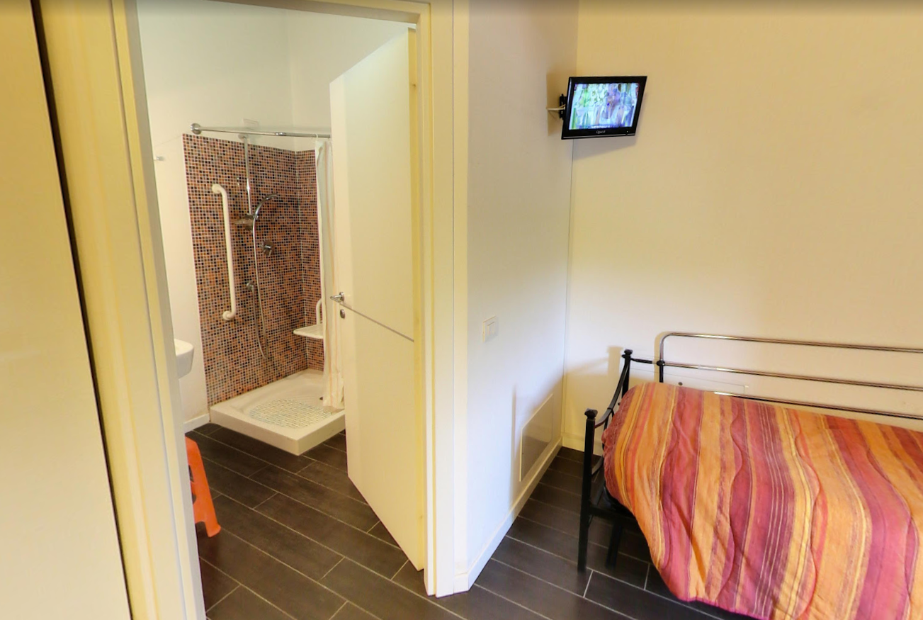 Camera da letto con letto singolo e bagno a vista. Una piccola TV è appesa alla parete.