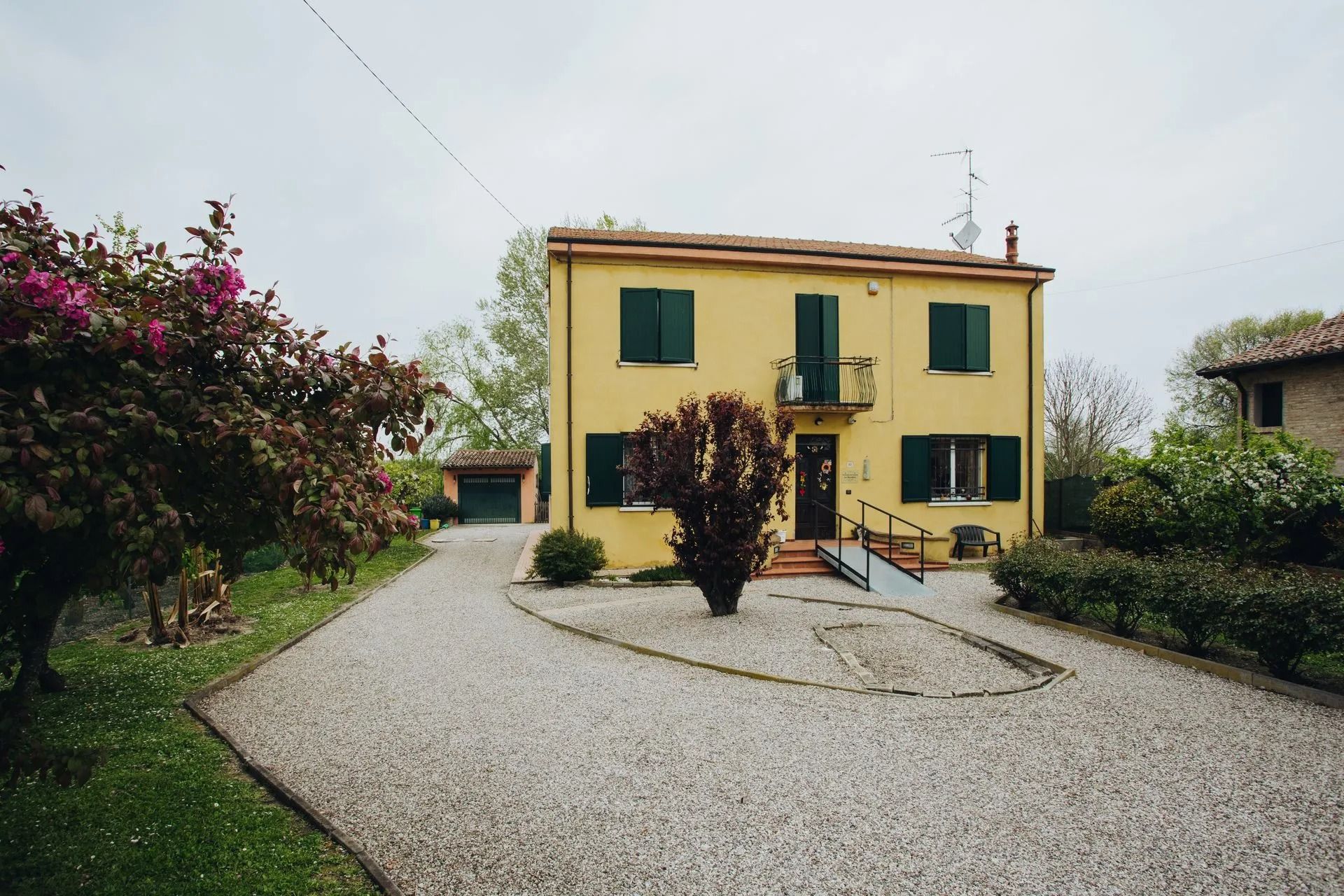 Casa gialla con vialetto di ghiaia e persiane verdi, circondata da alberi e vegetazione.