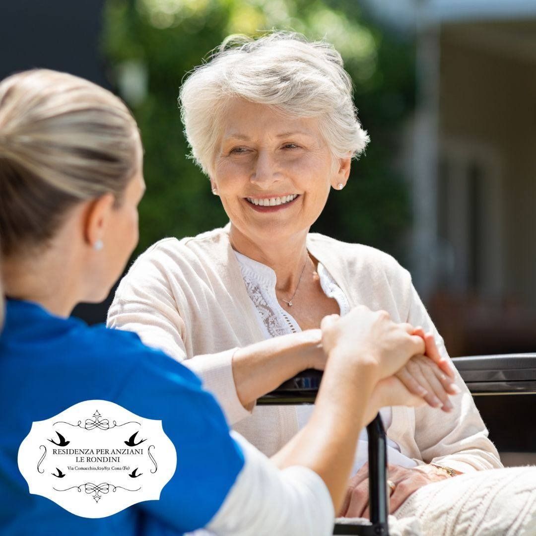 Un'assistente tiene per mano un anziano sorridente su una sedia a rotelle all'aperto.