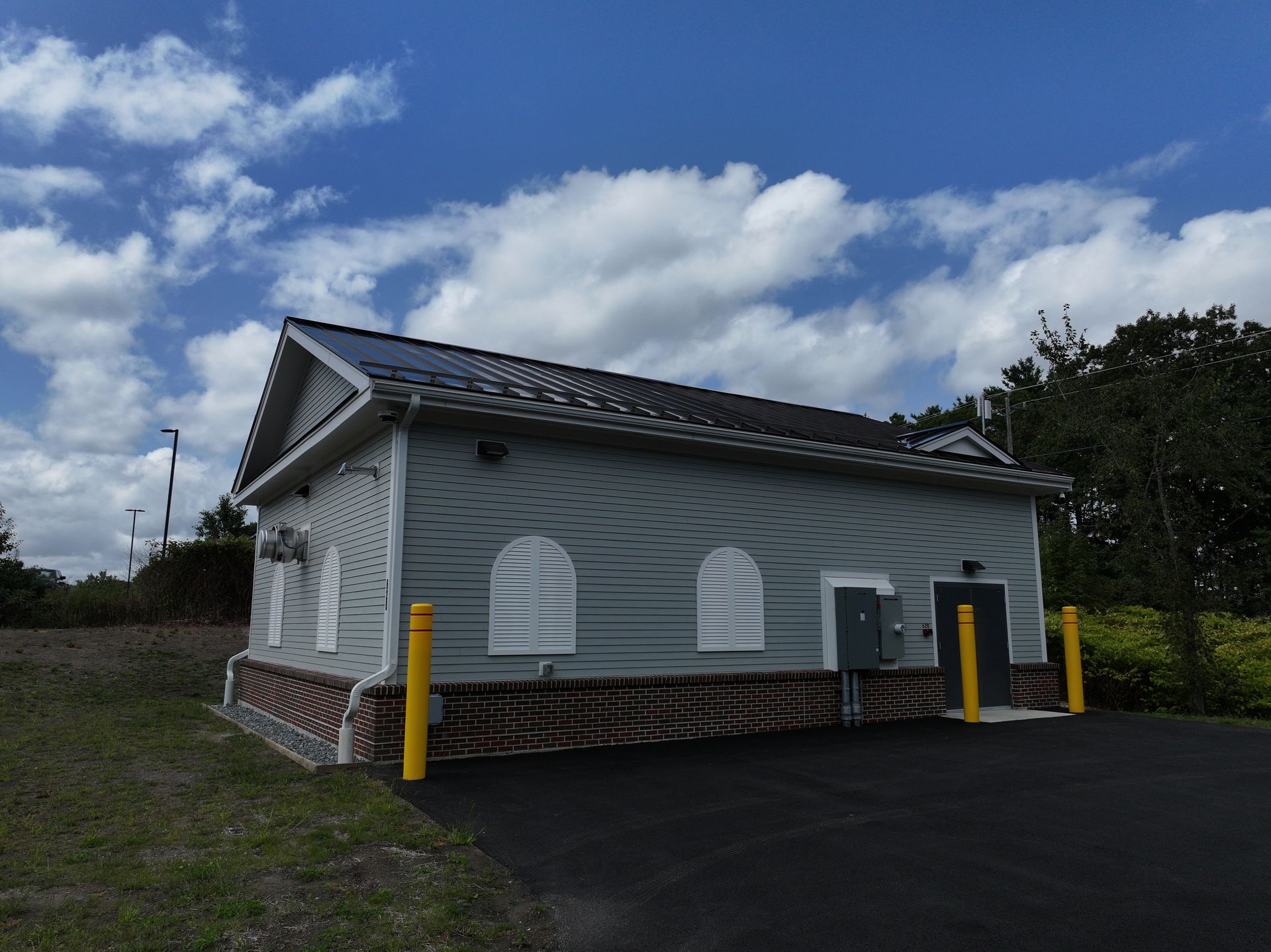gray water utility  building with shutters, dark roof, and yellow bollards on asphalt driveway