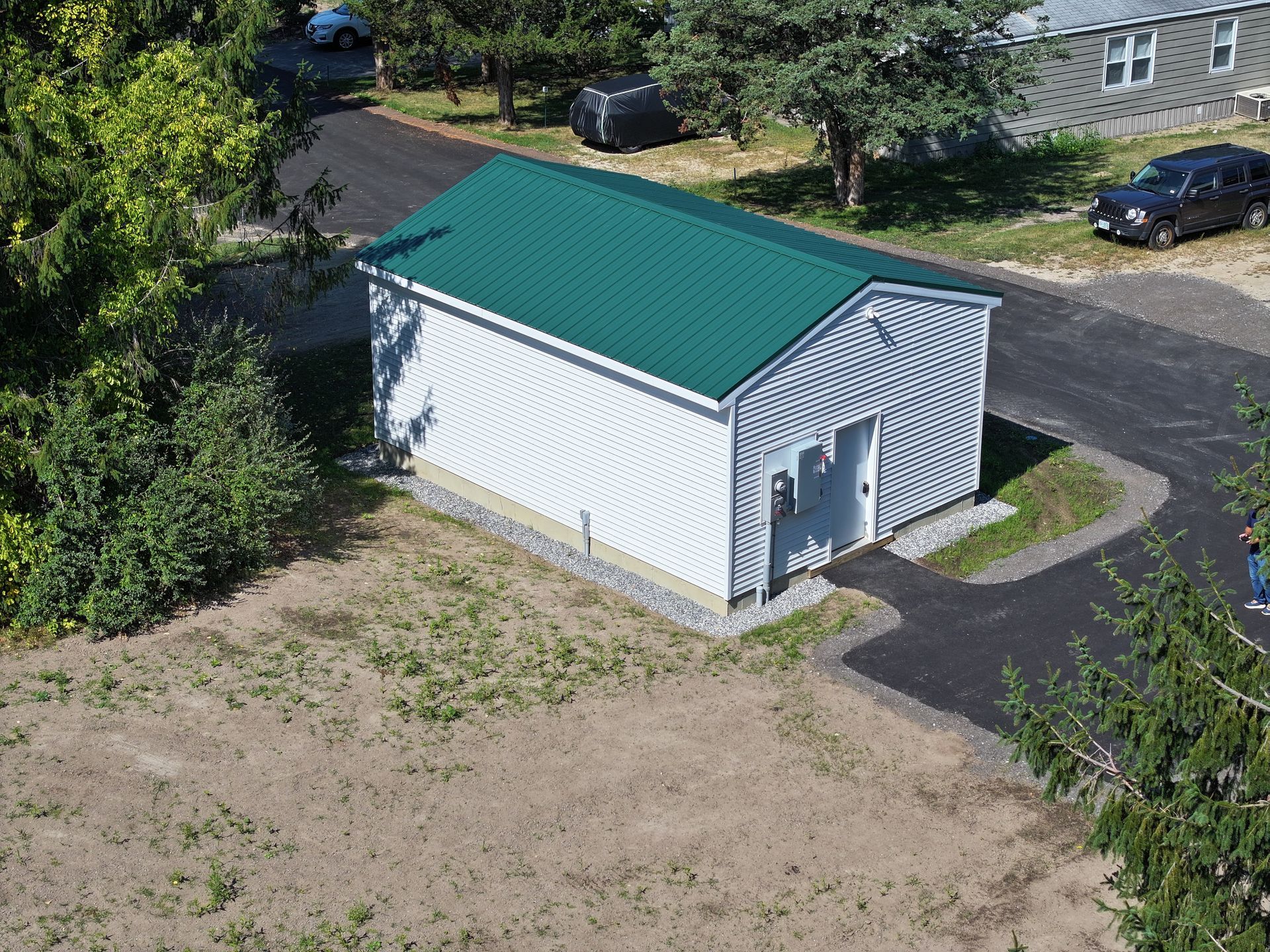 White garage with a green roof, door open. Asphalt driveway. Trees surround.