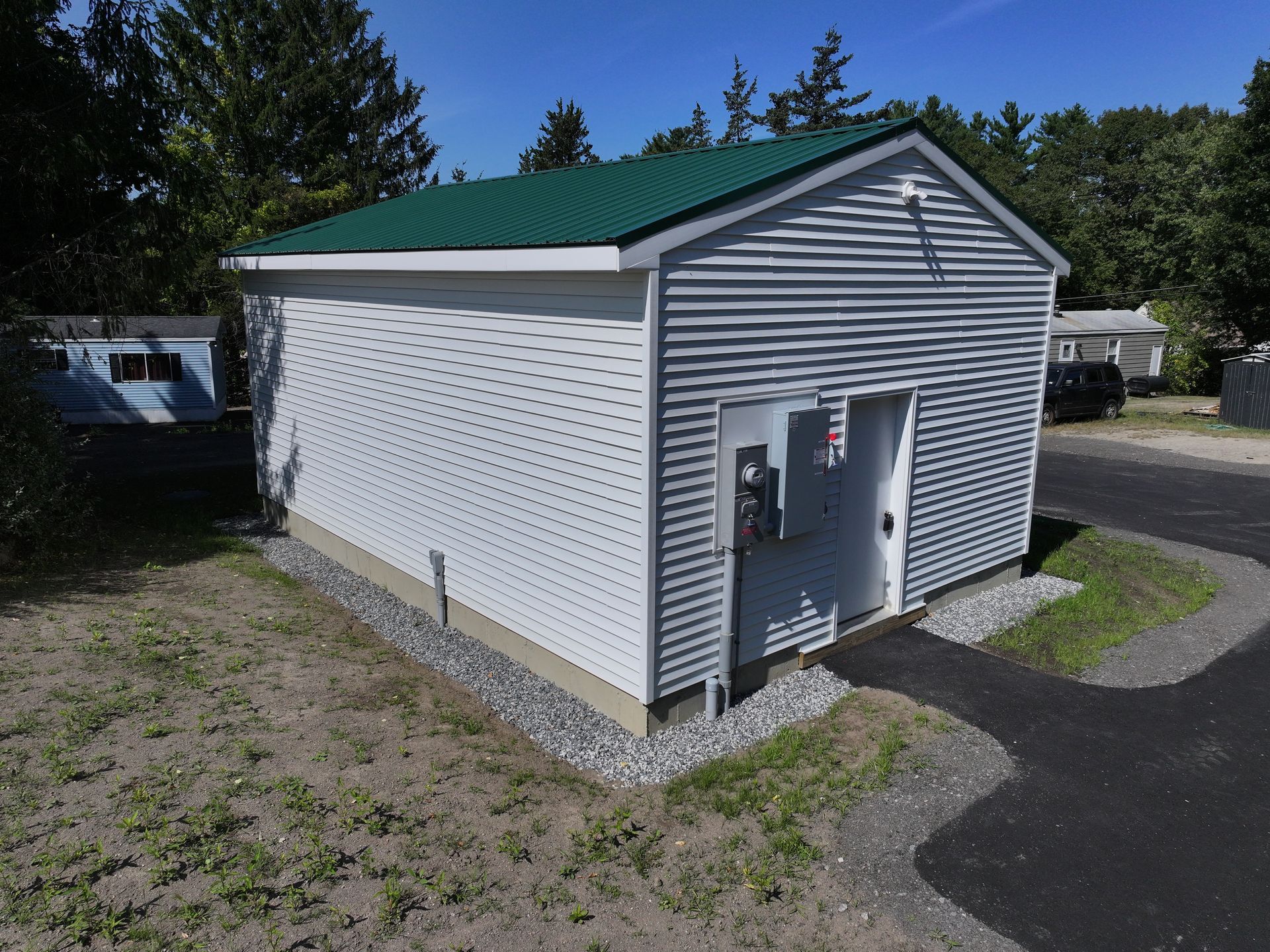 Small, white utility building with green roof, door, and gravel base, near a road.