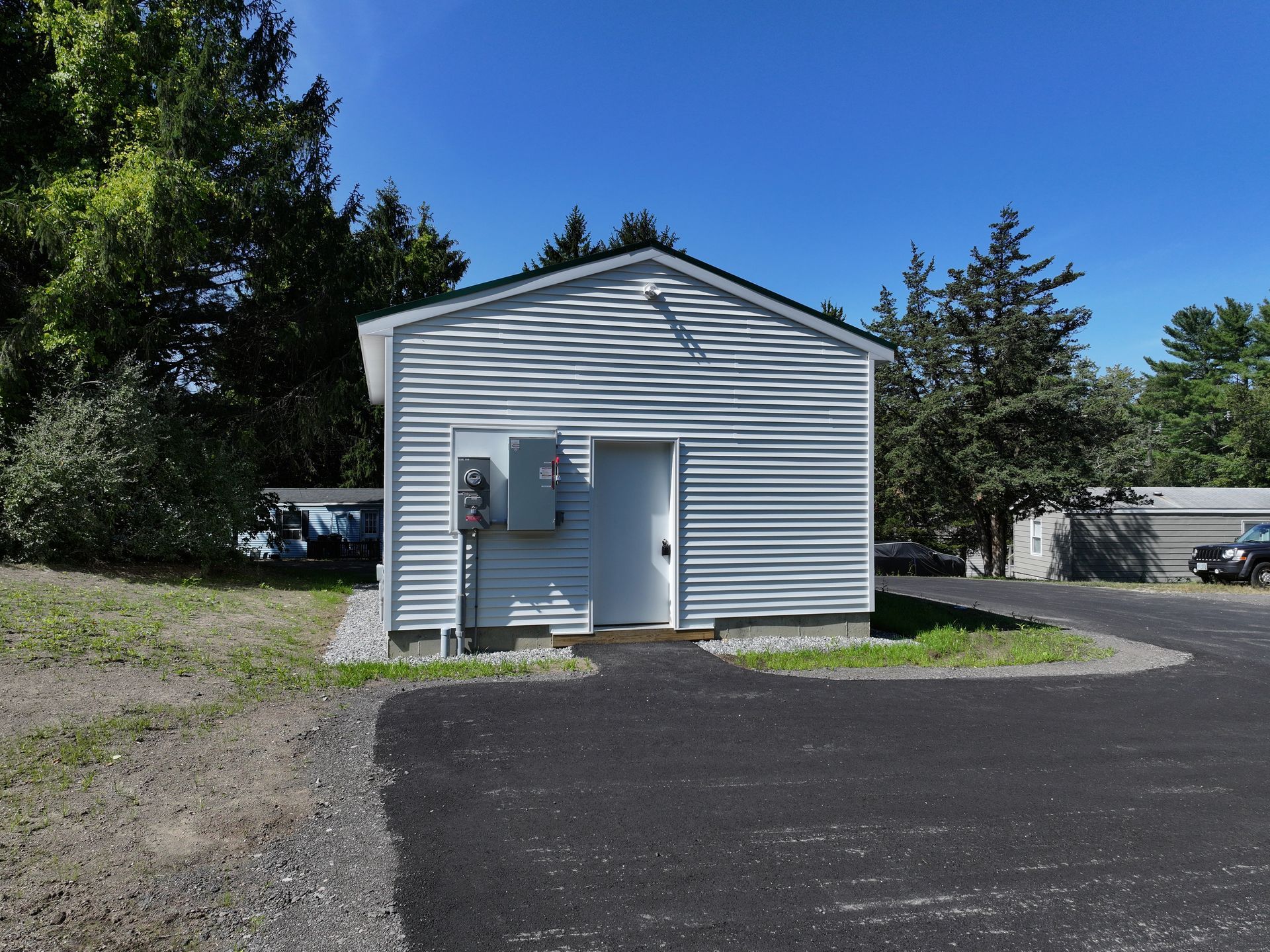 Small, light-blue sided building with a white door and electrical box, asphalt driveway, green grass, trees, and blue sky.