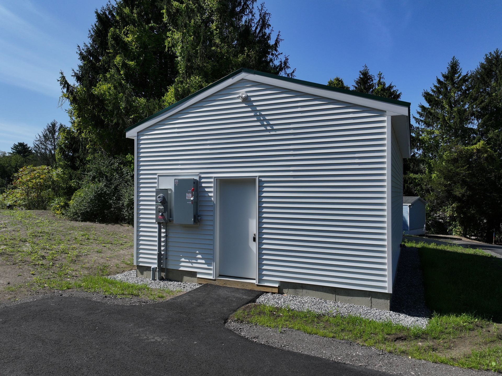White utility shed with electrical equipment, door, and asphalt path. Green trees in background.