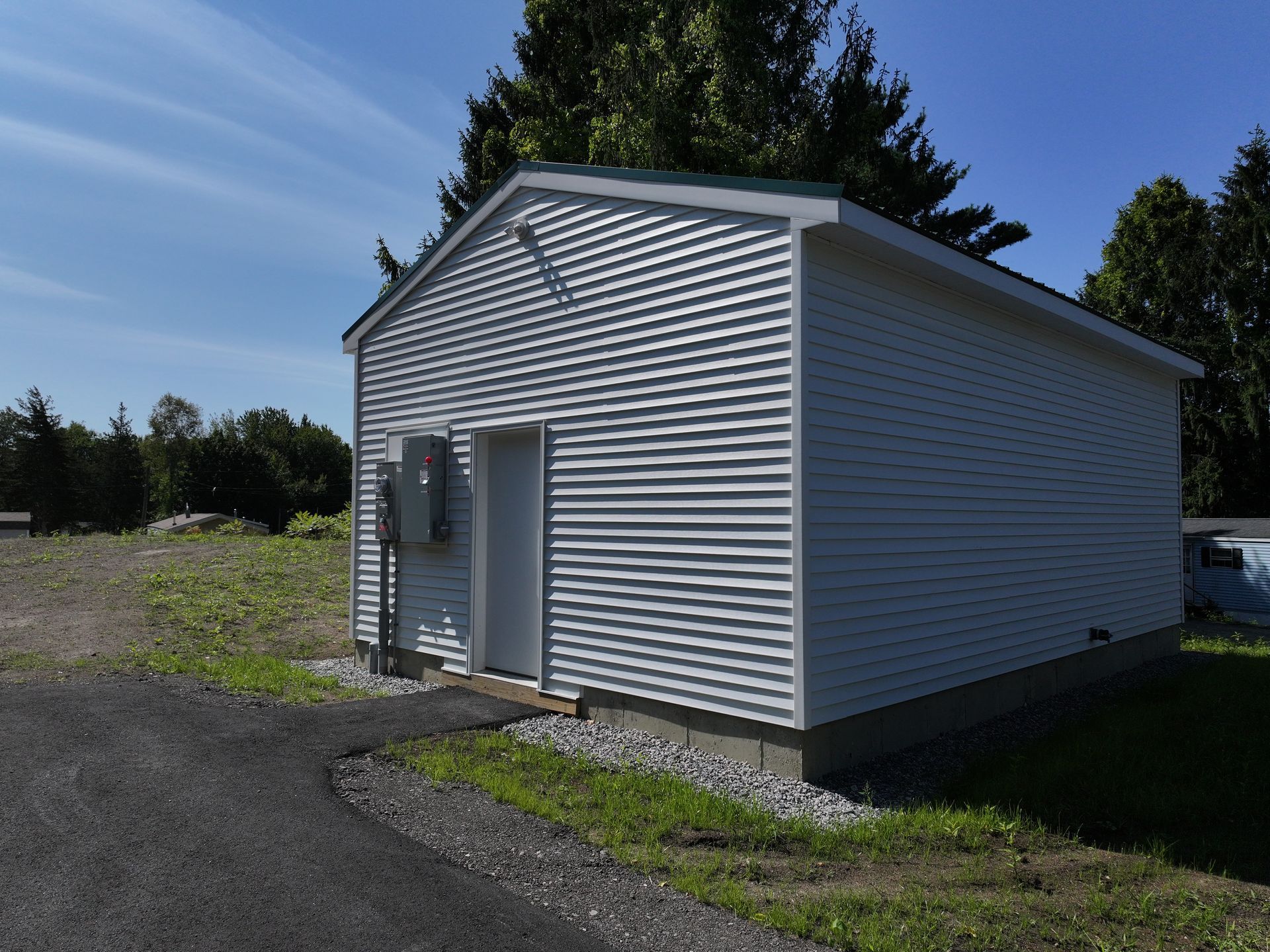 White sided shed with a green roof, door, and electrical box, on a concrete base, in a grassy area with a paved driveway.