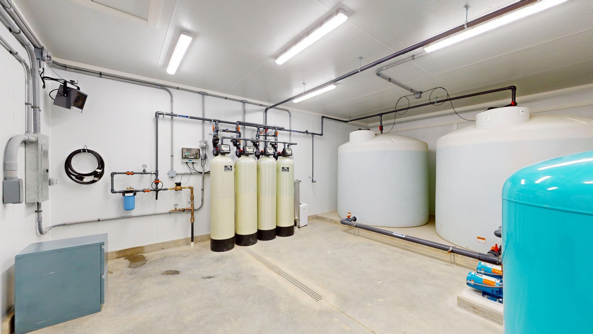 Water treatment room with tanks, filters, pipes, and control panel. White walls, concrete floor, overhead lighting.