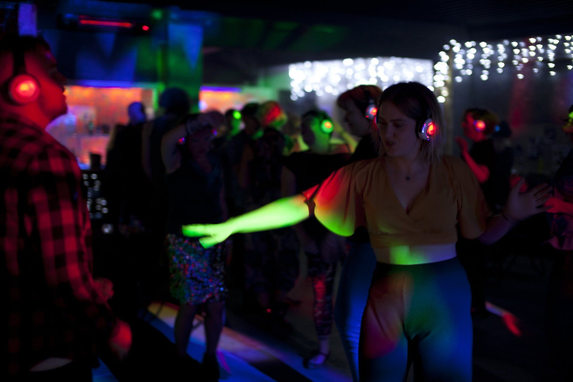 People dancing at a silent disco, wearing headphones with colorful lights. Low-lit club setting.