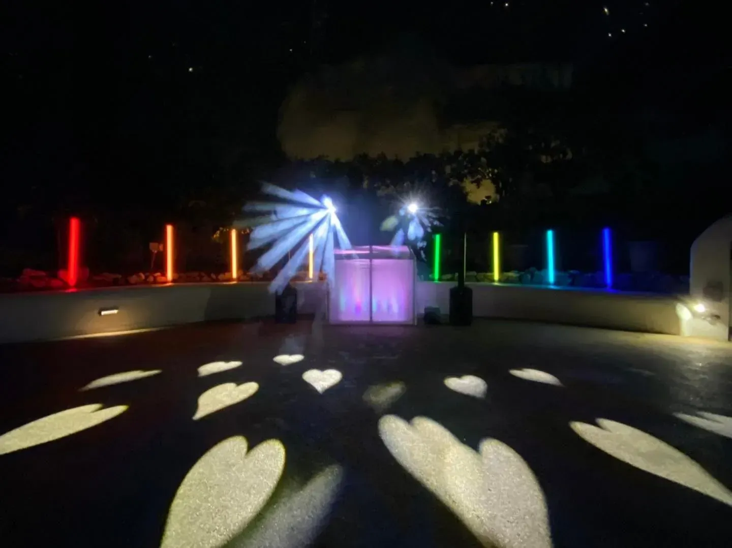 Nighttime outdoor event with DJ booth lit by spotlights, rainbow columns, and heart-shaped projections on the ground.