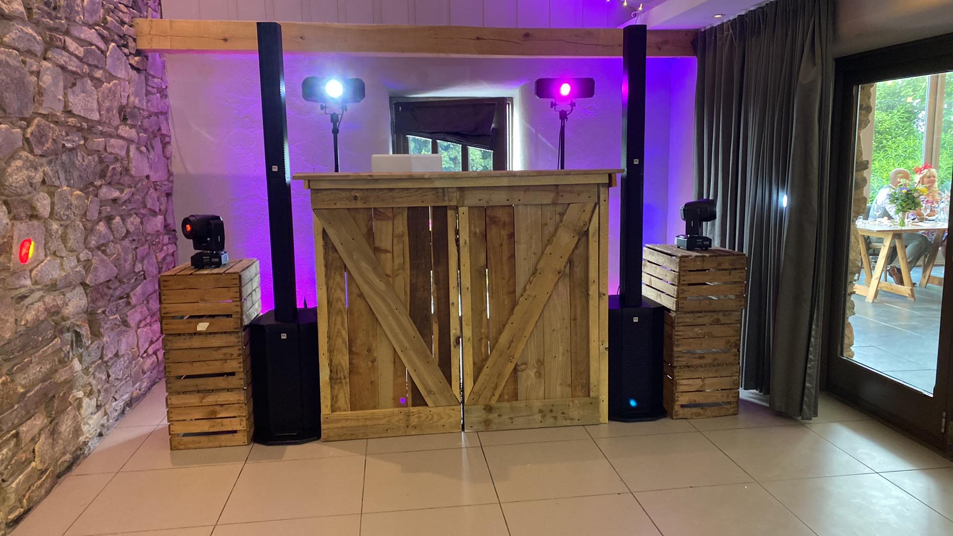 DJ setup with wooden crates, speakers, lights, and a wooden DJ booth, in a room with stone walls and a large window.