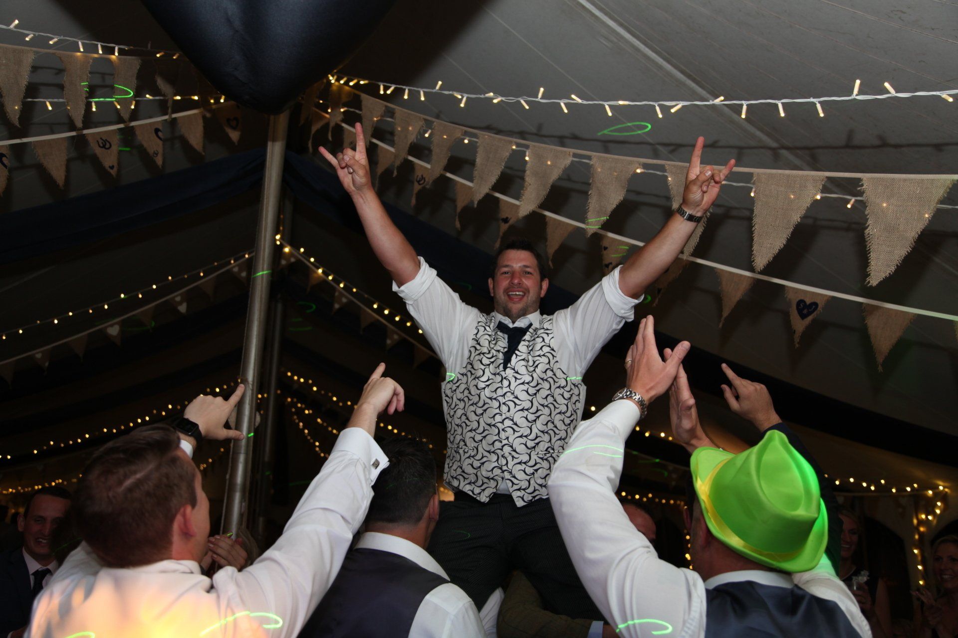 Man in vest on shoulders of others, arms raised in air, celebrating at party.
