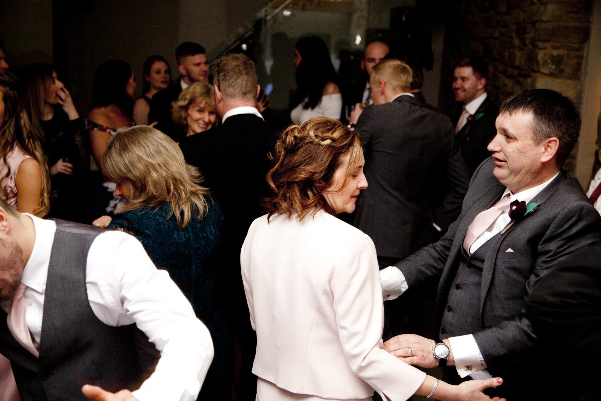 People dancing at a formal event. Men in suits, women in dresses, some smiling and enjoying the music. Dimly lit.